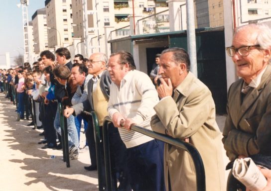 Grupo de hombres de pie en una fila, observando un evento en un entorno urbano.
